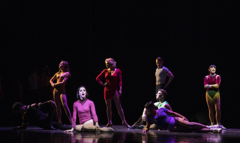 Performers of A Chorus Line pose on stage. Some stand with their arms crossed or on their hips. Other sing out to the audience while sitting on the floor.